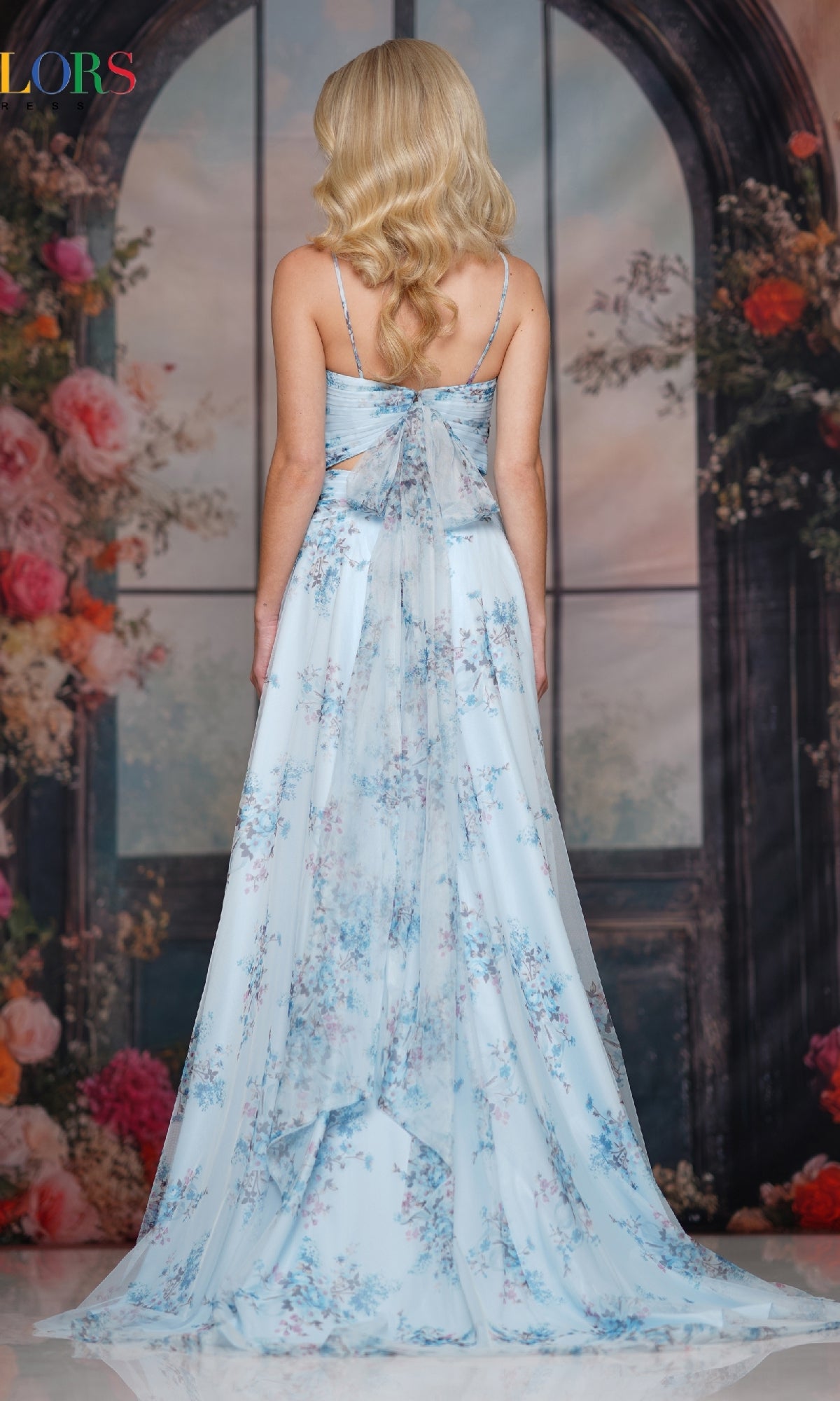A woman with long blonde hair stands facing away in front of arched windows and a floral backdrop, wearing the Cut-Out Long Print Prom Dress: Colors Dress 3813.